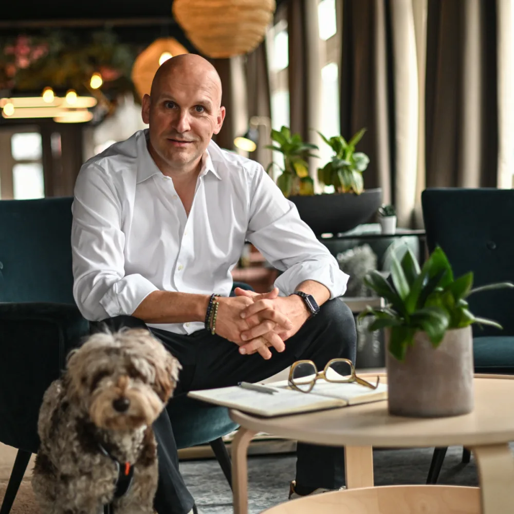 Marco Grüter sitzt in einer Lounge, die Hände gefaltet, sein Hund neben ihm